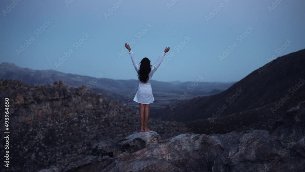 A woman wearing white dress standing on a cliff with hands up. A woman wearing white dress standing on a cliff with hands up.