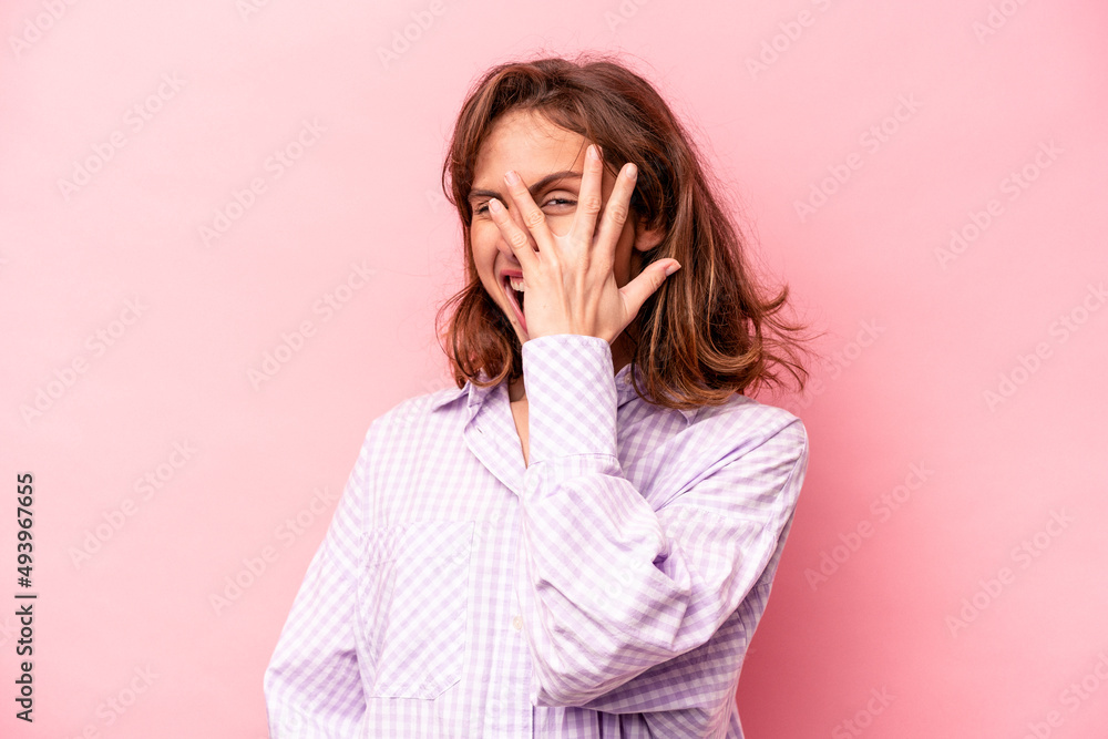 Young caucasian woman isolated on pink background blink at the camera through fingers, embarrassed covering face. Young caucasian woman isolated on pink background blink at the camera through fingers, embarrassed covering face.