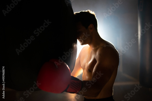 Tired boxer leaning on punching bag. Sweaty shirtless man in boxing gloves standing in gym against light. Sport and endurance concept photo