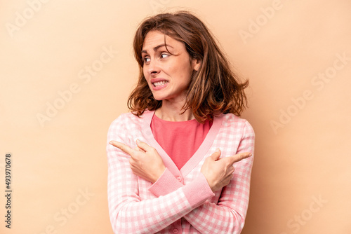 Young caucasian woman isolated on beige background points sideways, is trying to choose between two options.