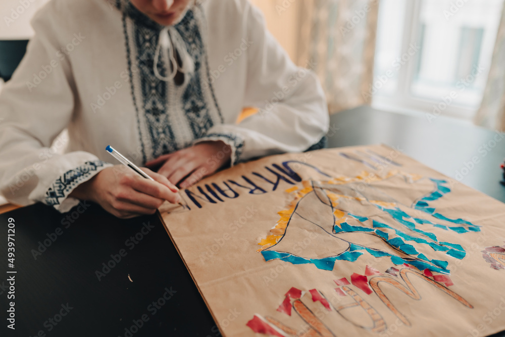 Obraz premium child in national ethnic embroidered white shirt draws poster in support of Ukraine, writes slogan against Russia's military actions