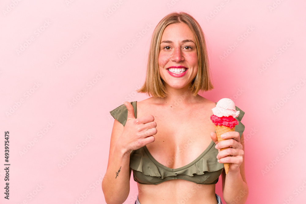 Young caucasian woman wearing a bikini and holding an ice cream isolated on pink background smiling and raising thumb up Young caucasian woman wearing a bikini and holding an ice cream isolated on pink background smiling and raising thumb up
