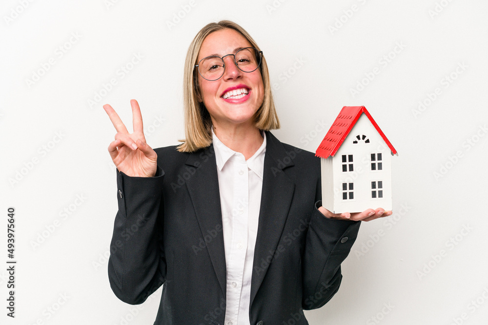 Young business caucasian woman holding a toy house isolated on white background joyful and carefree showing a peace symbol with fingers. Young business caucasian woman holding a toy house isolated on white background joyful and carefree showing a peace symbol with fingers.