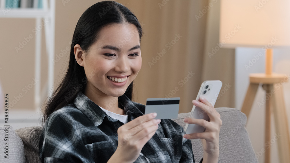 Smiling asian ethnic woman customer user holding bank credit card with virtual money typing smartphone at home. Happy multiethnic female shopper using easy mobile payments app making purchase online Smiling asian ethnic woman customer user holding bank credit card with virtual money typing smartphone at home. Happy multiethnic female shopper using easy mobile payments app making purchase online