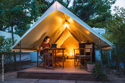Cozy open glamping tent with light inside and a woman using a laptop during dusk. Luxury camping tent for outdoor summer lifestyle concept photo
