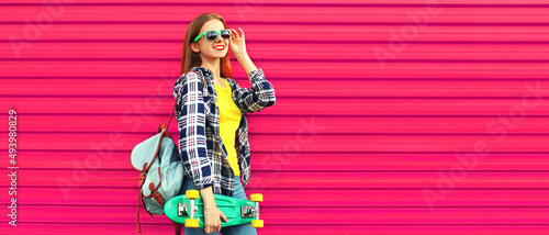 Portrait of stylish teenager girl with skateboard looking away wearing shirt, backpack on vivid pink background, blank copy space for advertising text © rohappy
