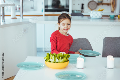 Little girtl helping mother to set the table for dinner, carrying the plates, kitchen interior, cropped, copy space. Smiling girl setting table.