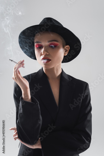 woman in black fedora hat and blazer smoking isolated on grey.