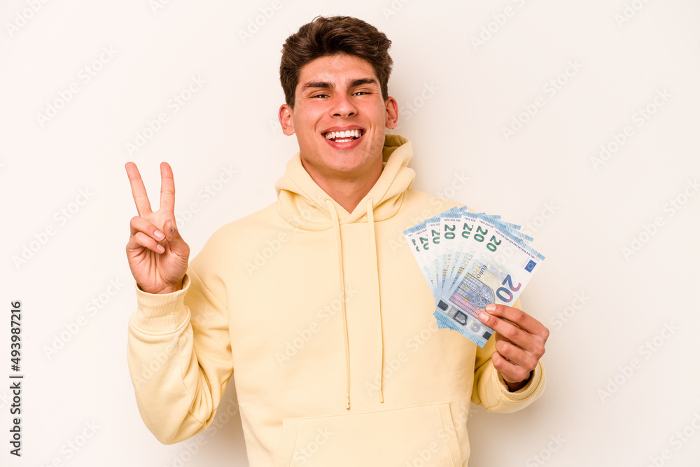 Young caucasian man holding banknotes isolated on white background joyful and carefree showing a peace symbol with fingers. Young caucasian man holding banknotes isolated on white background joyful and carefree showing a peace symbol with fingers.