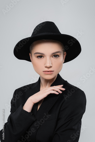 charming woman in black blazer and brim hat looking at camera isolated on grey.