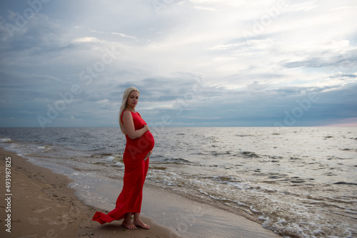 Beautiful Latvian pregnant woman in red dress standing at the beach during sunset