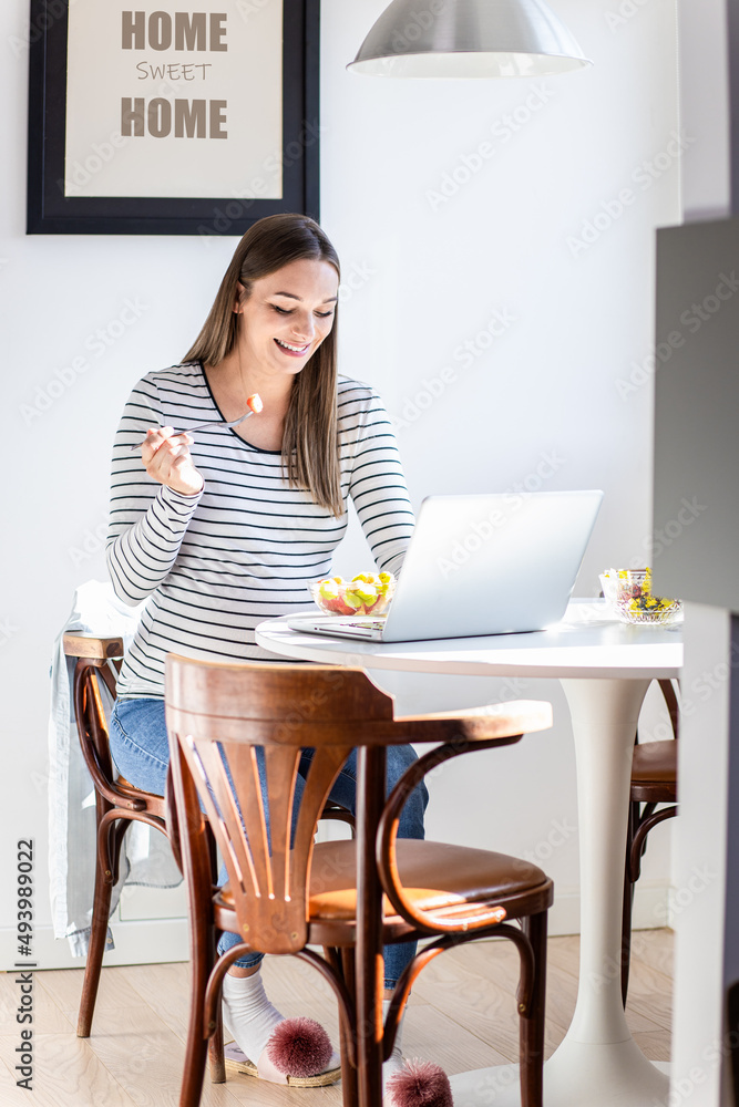 Pregnant woman sitting at table at home using laptop for video call.