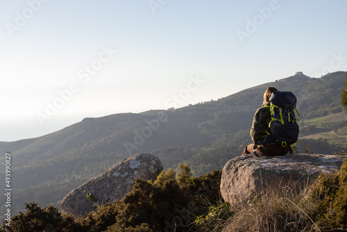 Rear view of young backpacker sitting on stone looking at view. Woman with backpack resting on top of mountain. Hiking concept