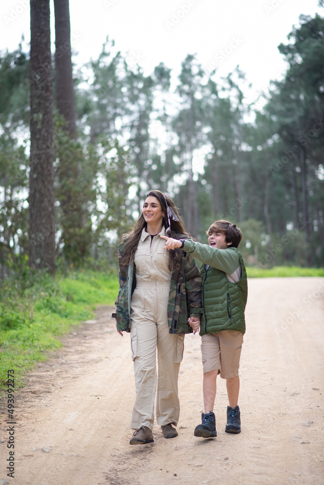 Satisfied mother and son walking in forest. Dark-haired wo and boy in coats walking on cloudy day, pointing. Family, nature, leisure concept Satisfied mother and son walking in forest. Dark-haired wo and boy in coats walking on cloudy day, pointing. Family, nature, leisure concept