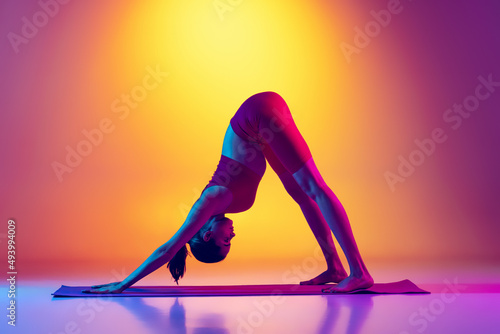 Portraity of young sportive girl doing stretching exercises isolated over gradient pink and yellow background in neon