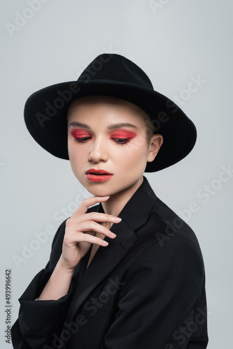 pretty woman in black brim hat and blazer touching chin isolated on grey.