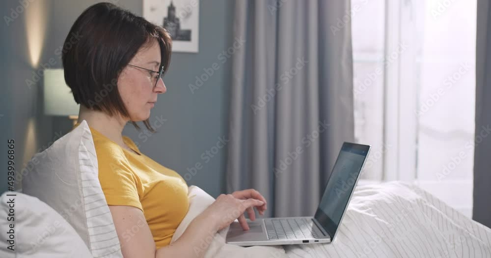 Young caucasian female in glasses, scrolling and typing on computer keyboard in bedroom. Pretty woman lying in the bed and using laptop notebook to work. Young caucasian female in glasses, scrolling and typing on computer keyboard in bedroom. Pretty woman lying in the bed and using laptop notebook to work.
