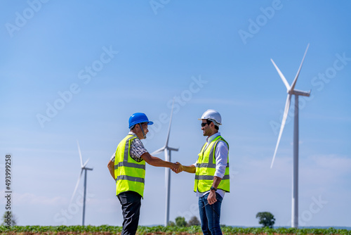 Engineer wearing uniform ,helmet show handshake corporate work in wind turbine farms rotation to generate electricity energy. Green ecological power energy generation wind sustainable energy concept.