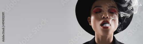 woman in black brim hat, with bright makeup, exhaling smoke isolated on grey, banner.