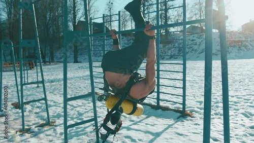 Bodybuilder with a naked torso with a drager balloon on the back is training in a winter park and doing the horizon exercise on the crossbar photo