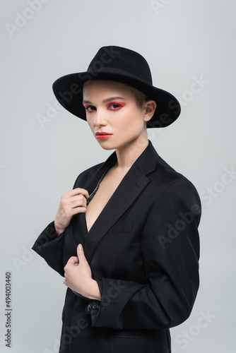 sensual young woman in brim hat touching black blazer while looking at camera isolated on grey.