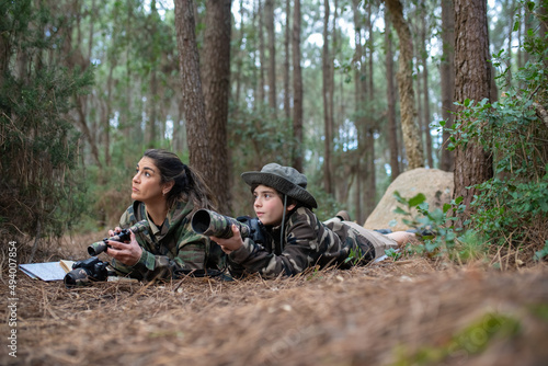 Attentive mother and son taking pictures in forest. Family with modern cameras lying on ground, holding camera and binoculars. Parenting, family, leisure concept