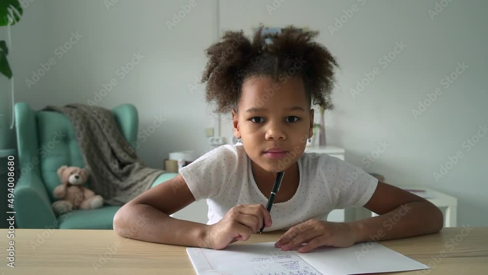 Little girl speaking and writing at table in front of device in home interior spbd. Portrait of American African child looks with smile and speaks, writes in notebook and sits at desk in light room Little girl speaking and writing at table in front of device in home interior spbd. Portrait of American African child looks with smile and speaks, writes in notebook and sits at desk in light room