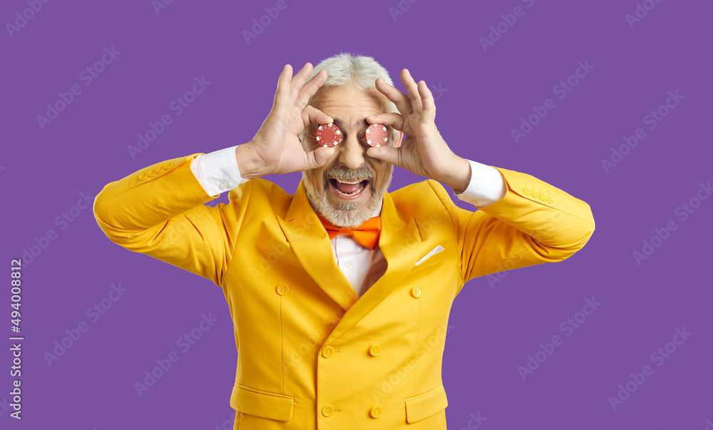 Studio shot of happy funny goofy bearded adult man in funky yellow suit standing isolated on purple background, covering both his eyes with two poker chips and laughing. Casino, luck, fun concept Studio shot of happy funny goofy bearded adult man in funky yellow suit standing isolated on purple background, covering both his eyes with two poker chips and laughing. Casino, luck, fun concept