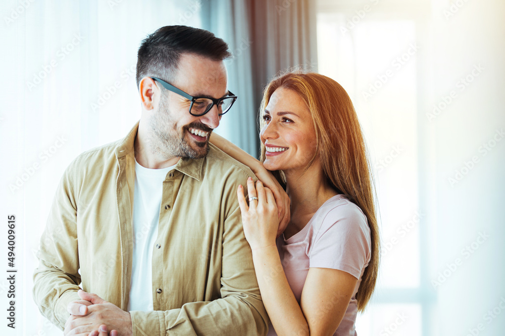 Smiling couple hugging each other and standing near window while looking outside. Happy and romantic man embracing hispanic wife from behind while standing at home with copy space. Smiling couple hugging each other and standing near window while looking outside. Happy and romantic man embracing hispanic wife from behind while standing at home with copy space.