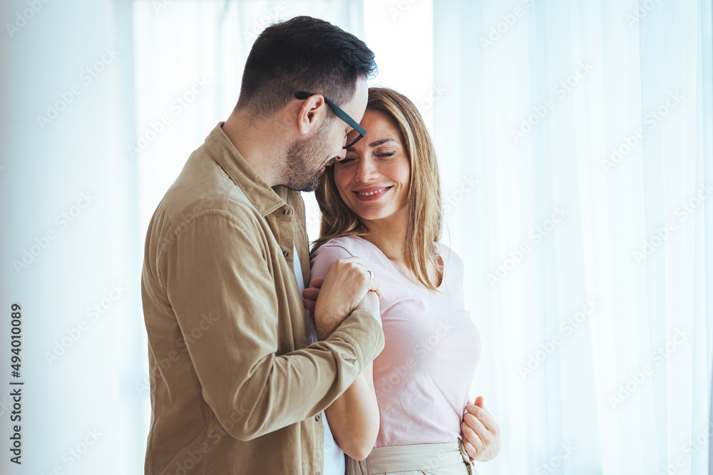 Smiling couple hugging each other and standing near window while looking outside. Happy and romantic man embracing hispanic wife from behind while standing at home with copy space. Smiling couple hugging each other and standing near window while looking outside. Happy and romantic man embracing hispanic wife from behind while standing at home with copy space.
