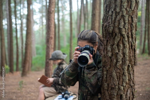 Absorbed woman with camera in forest. Mother in coat with ponytail taking pictures. Blurry son in background. Nature, leisure concept
