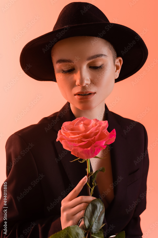 Fototapeta premium charming woman in black brim hat holding pink rose on coral background.