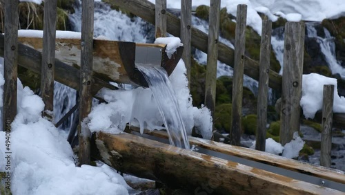 Clear fast-flowing meltwater between wooden beams with the partly snow-covered Kyogoku waterfalls in Fukidashi Park in Japan in the background behind the wooden fence. Close up shot photo