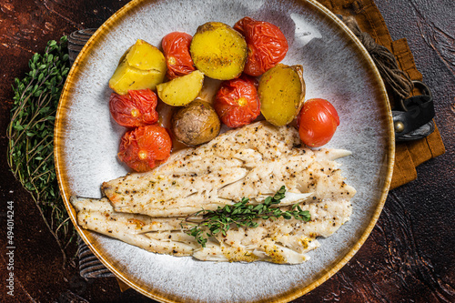 Roasted Pollock fish fillet in plate with garnish tomato and potato. Dark background. Top view