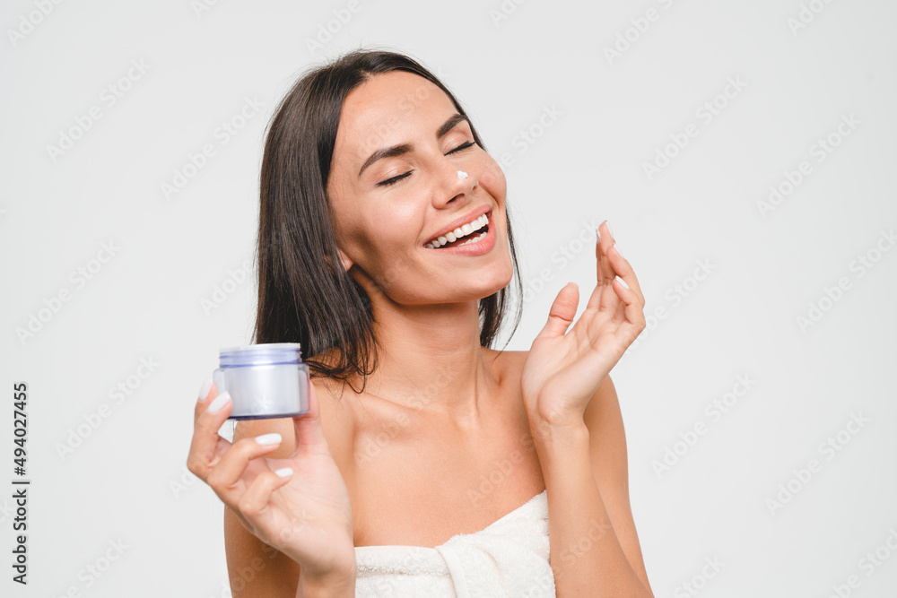 Happy caucasian young woman applying moisturizer on her face, face skin care with soft mask cream creme, rejuvenation anti-acne anti-aging effect isolated in grey background. Beauty concept Happy caucasian young woman applying moisturizer on her face, face skin care with soft mask cream creme, rejuvenation anti-acne anti-aging effect isolated in grey background. Beauty concept