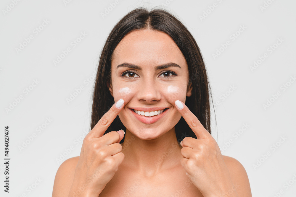 Cropped portrait of a young girl woman using softening moisturizing face cream for skin face care. Beauty treatment. Woman applying cosmetics on her face isolated in grey background Cropped portrait of a young girl woman using softening moisturizing face cream for skin face care. Beauty treatment. Woman applying cosmetics on her face isolated in grey background