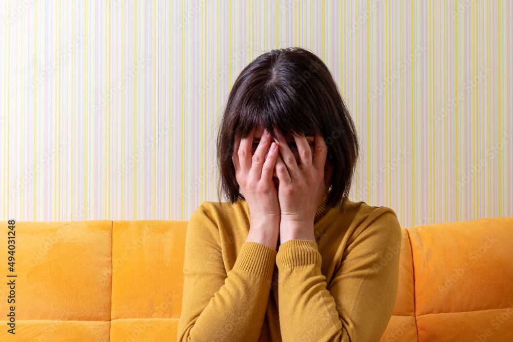 Upset crying middle aged woman suffering from stress covering her face with her hands on an orange sofa. Middle age crisis. Mental health concept. Upset crying middle aged woman suffering from stress covering her face with her hands on an orange sofa. Middle age crisis. Mental health concept.