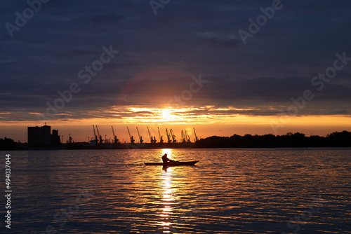 Alone kayaker under a colorful sky at sunset on a Danube river against the backdrop of port cranes