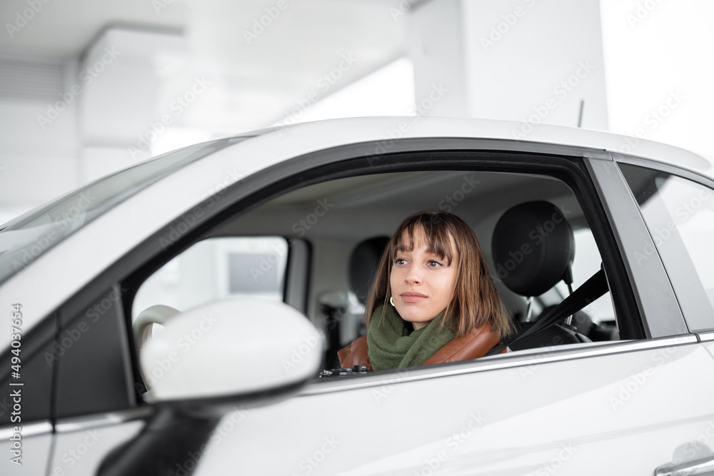 Portrait of a confident young woman sitting on the driver's seat of her white car at gas station. Caucasian woman wearing winter clothes