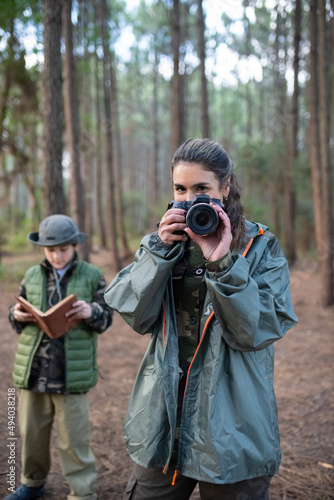 Attractive woman with camera in forest. Female model in sportive clothes smiling at camera. Blurry boy in background. Hobby, photography concept © KAMPUS