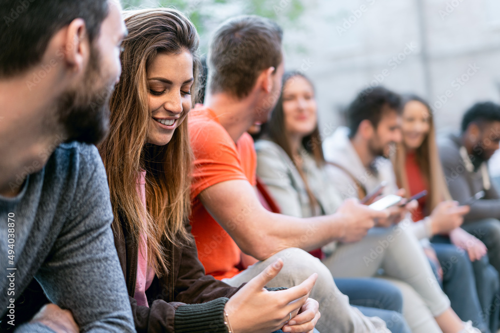 Naklejka premium Group of trendy young people chatting together sitting on a bench outdoors. Students having fun together. Focus on the blonde girl