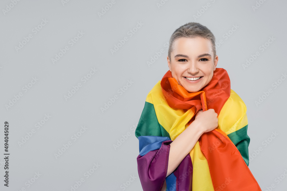 joyful woman looking at camera while wrapping in lgbt flag isolated on grey. joyful woman looking at camera while wrapping in lgbt flag isolated on grey.