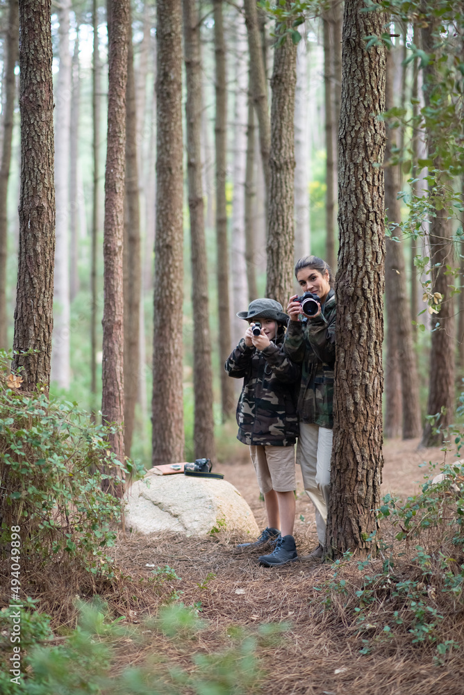 Mother and son spending time in forest. Woman and son in casual clothes with cameras peeking from behind trees. Hobby, family, nature, photography concept