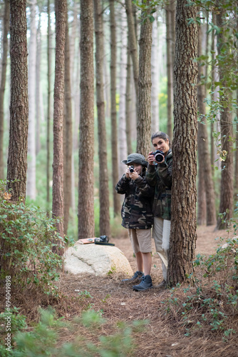Mother and son spending time in forest. Woman and son in casual clothes with cameras peeking from behind trees. Hobby, family, nature, photography concept