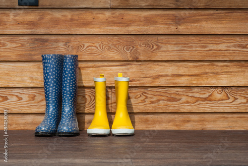 Blue and yellow rubber boots stand on the terrace. Adult and children's boots stand against the background of a wooden wall at home