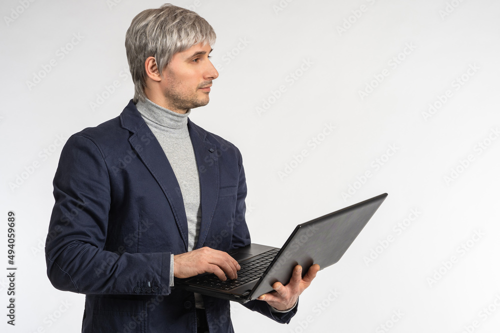 Successful man. Businessman with laptop. Man in business suit stands on gray. Male entrepreneur. Gray-haired guy is holding laptop. Copyspace. Businessman is typing text on notebook. IT specialist