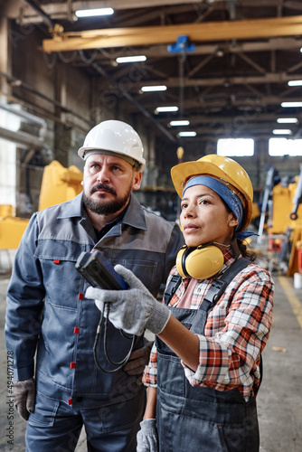 Young female technician in workear pressing button on remote control while regulating height of huge industrial machine photo