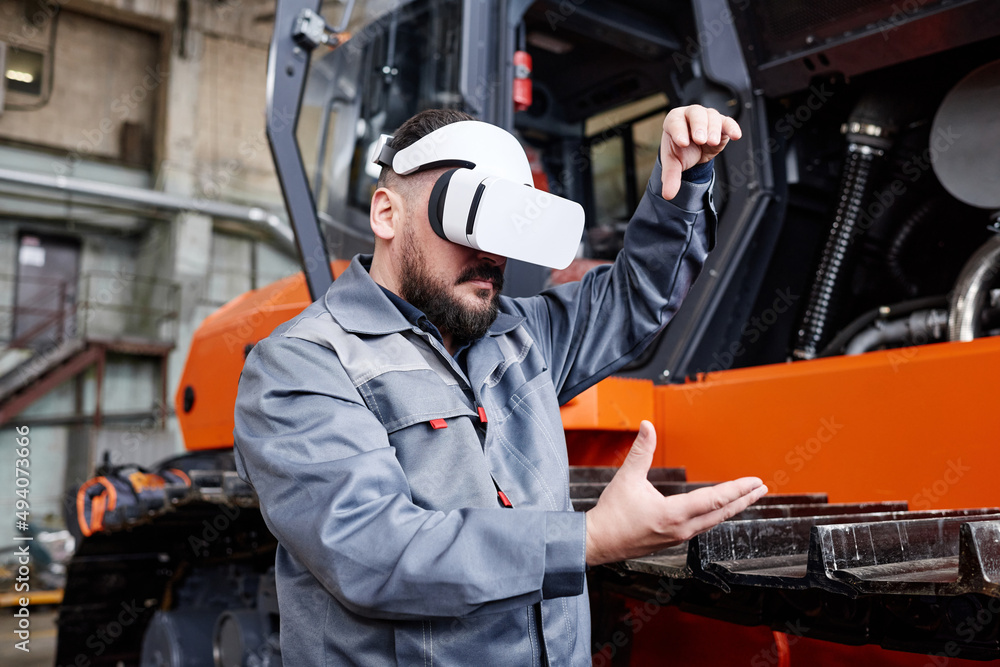 Contemporary engineer in vr headset forming shape of virtual machine by his hands while standing in warehouse or workshop