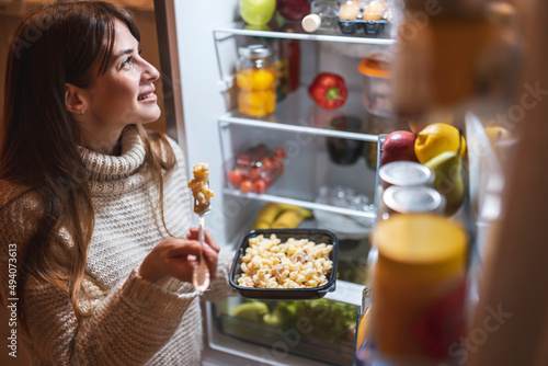 Woman standing by an open fridge eating late at night