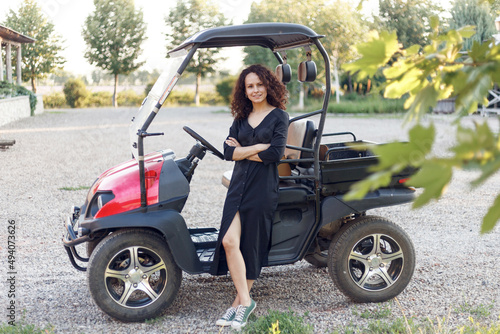 Cheerful woman with curly hair in black dress, poses near vehicle and smiling.